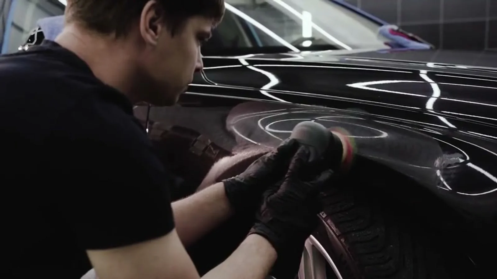 Machine polisher being worked across a dark front wing, hard overhead light revealing swirl marks and buffer trails in the paint