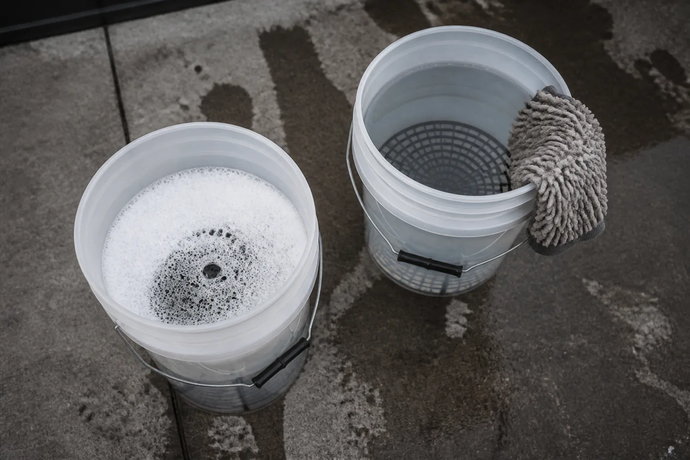 Two detailing buckets on wet concrete, one full of suds and one of rinse water, with a wash mitt draped over the rinse bucket rim
