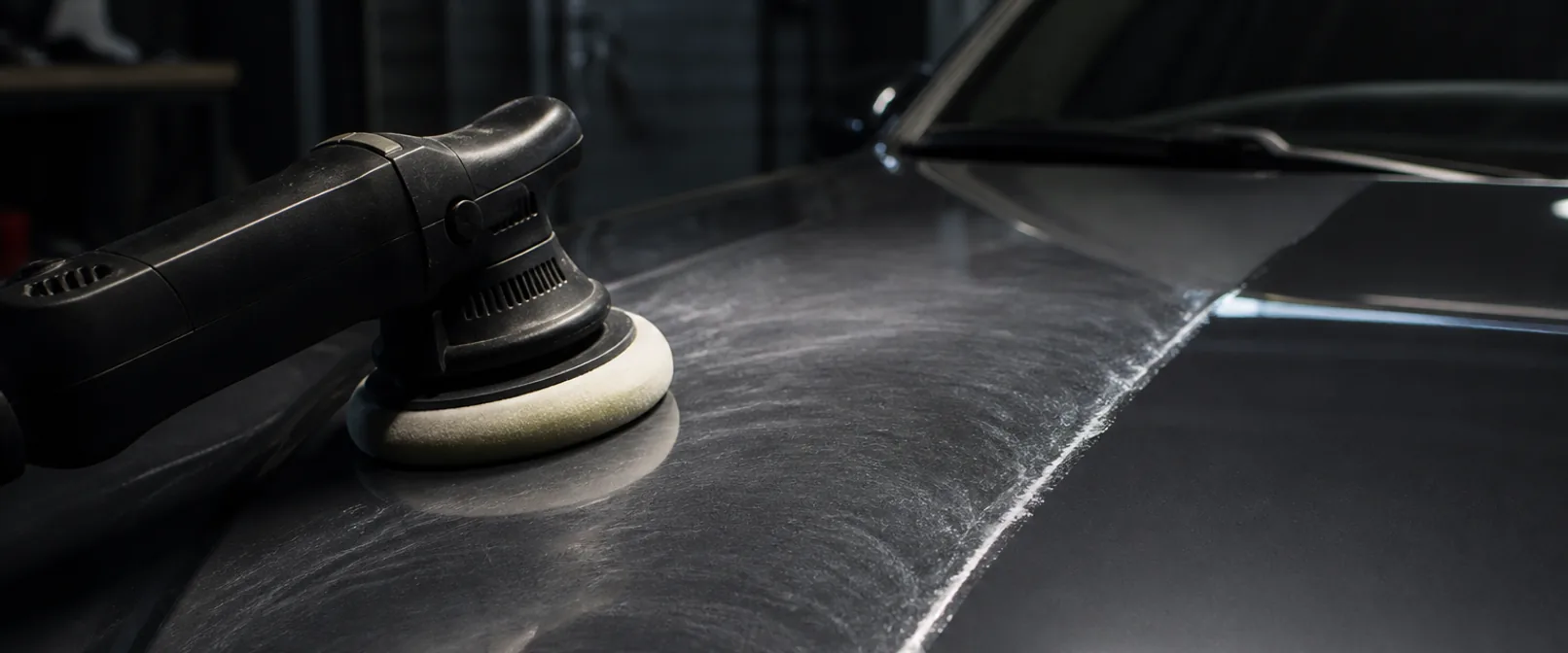 Dual-action polisher resting on a dark car bonnet at a correction line, the polished side holding a sharp reflection of the overhead light while the unpolished side scatters it into a grey haze of swirl marks